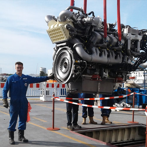 Ein Werftmitarbeiter hält großen Motor fest der am Kran hängt.